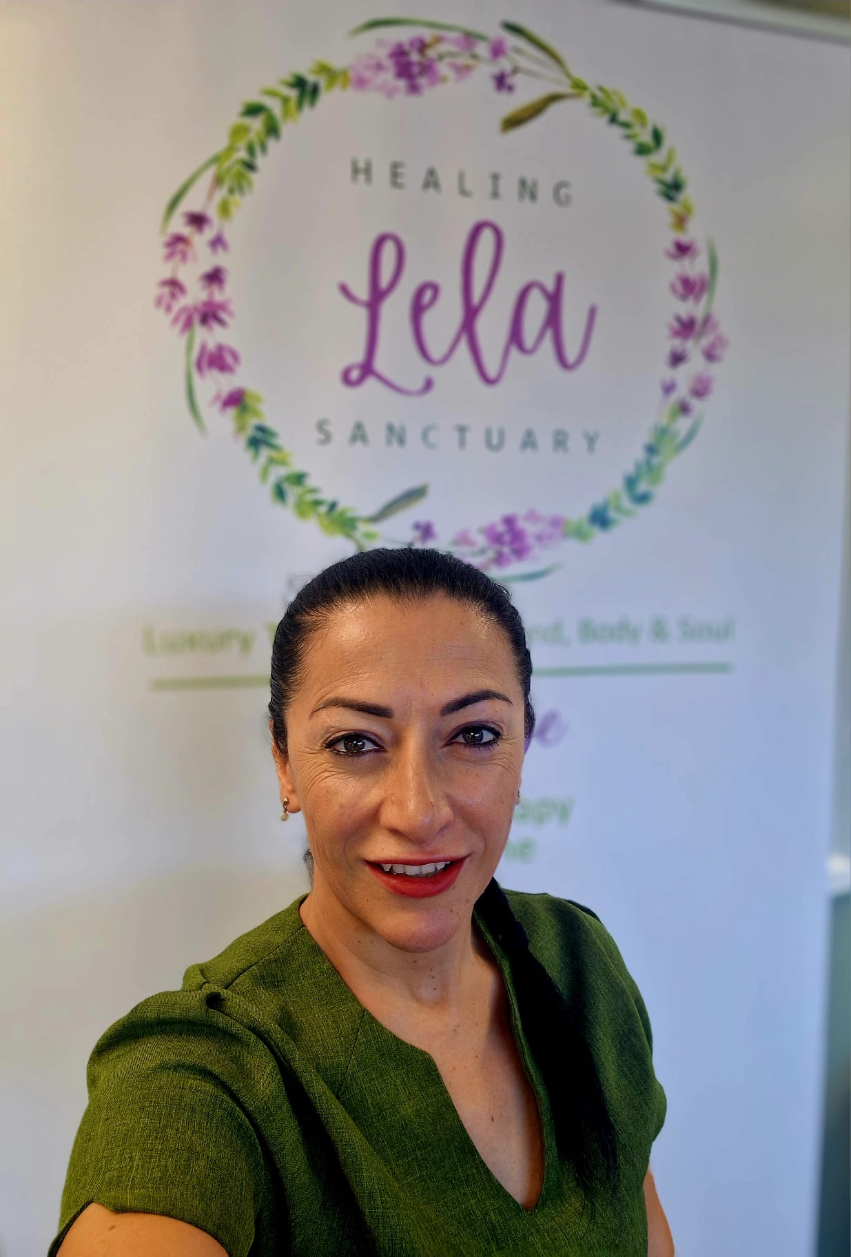 Aleks from Lela Healing Sanctuary, smiling, in a green therapist's tunic in front of her Lela Healing Sanctuary banner