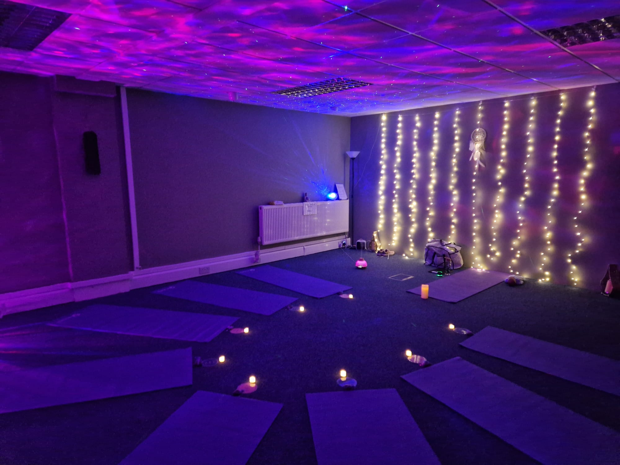 The community space set up for a class, with yoga mats in a circle, candles, a curtain of fairy lights and soft purple lighting
