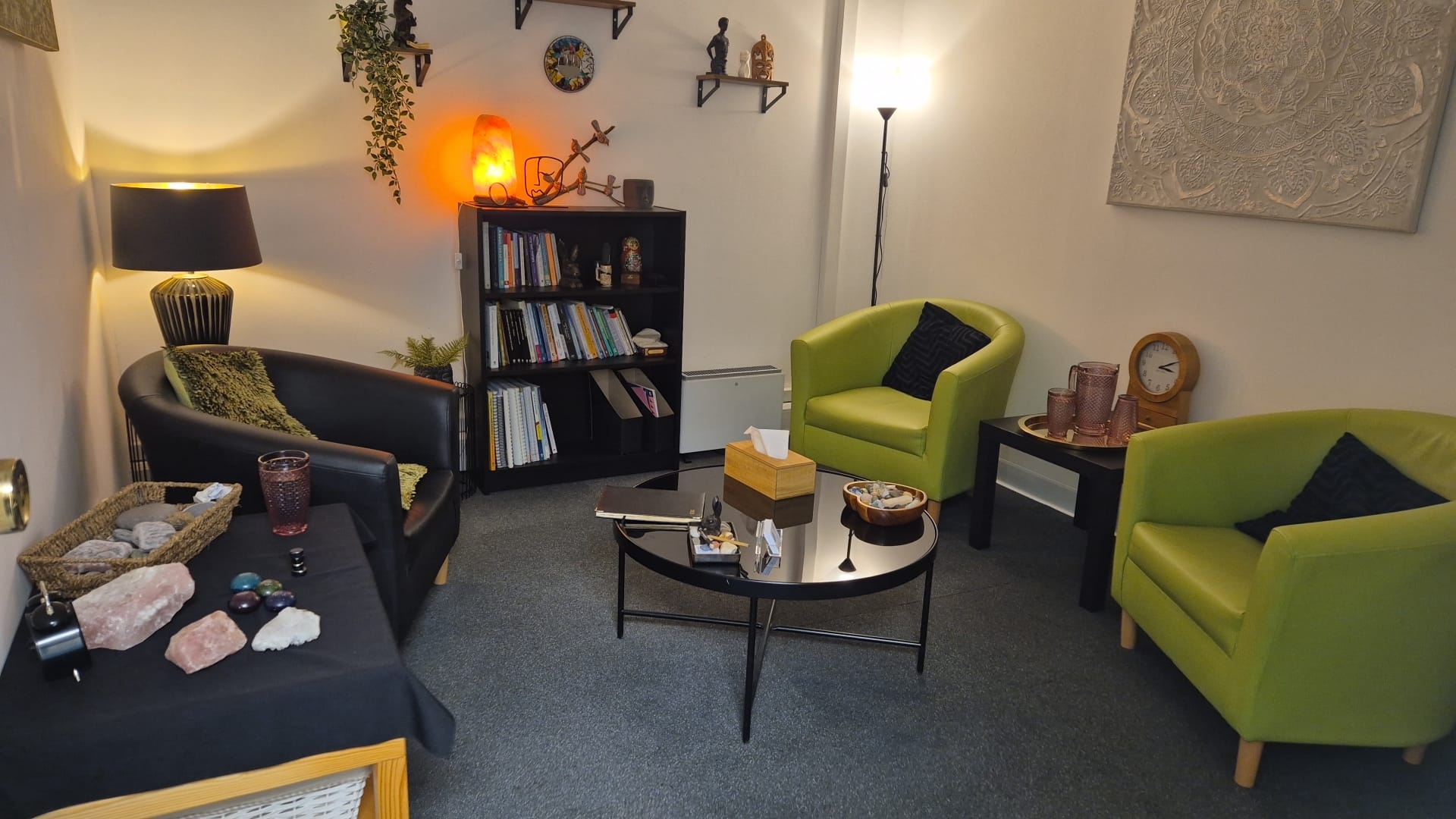 A cosy therapy room with green armchairs, a coffee table, a bookshelf and soft lamp light