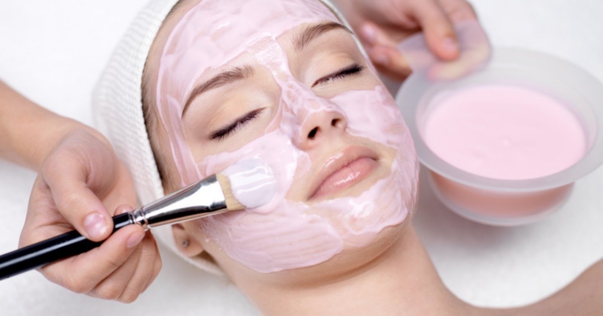 A therapist brushing a pink face mask onto a relaxed client during a facial treatment