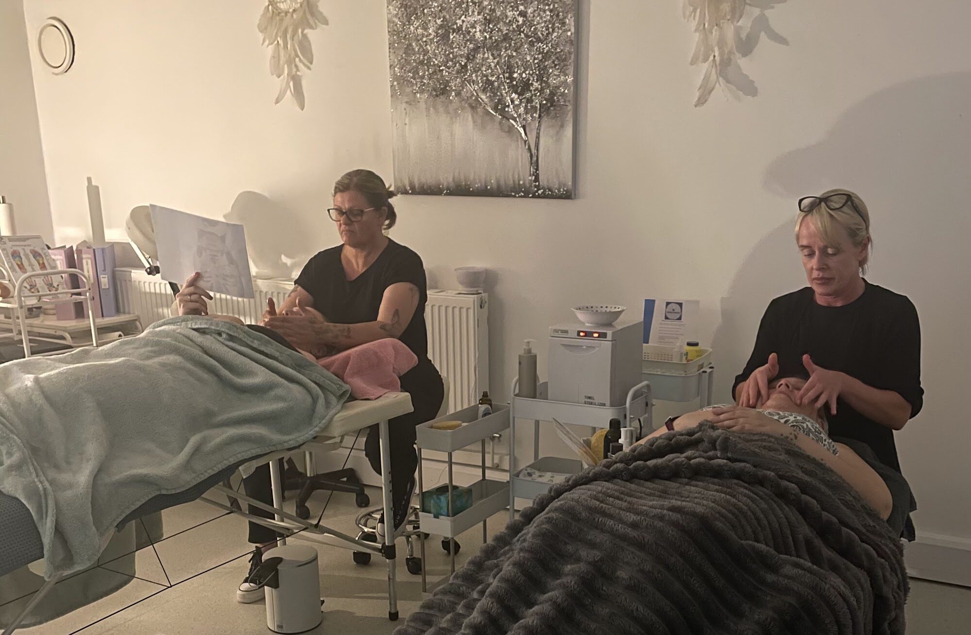 Two students practising facial treatments on clients lying on treatment couches during a facial training day