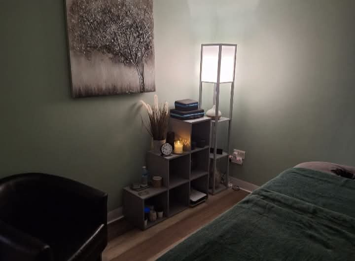A calm treatment room with sage green walls, a massage couch, a tall lamp and candles on the shelves