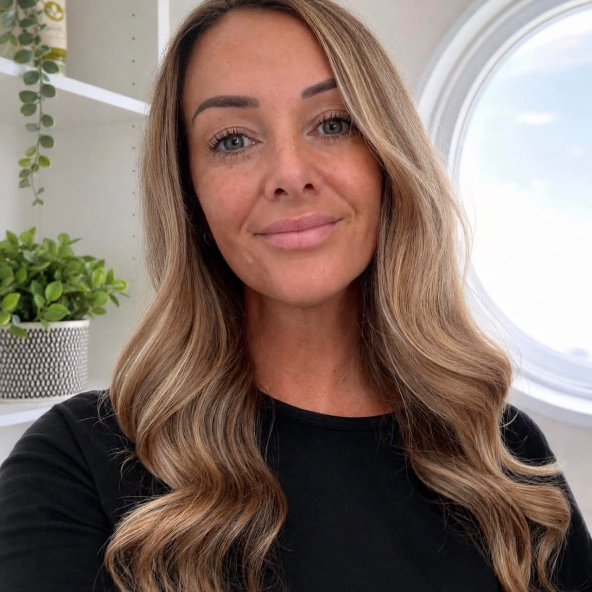 Lianne from Brows and Beauty by Lianne, smiling, in the bright treatment room with a round porthole window behind her