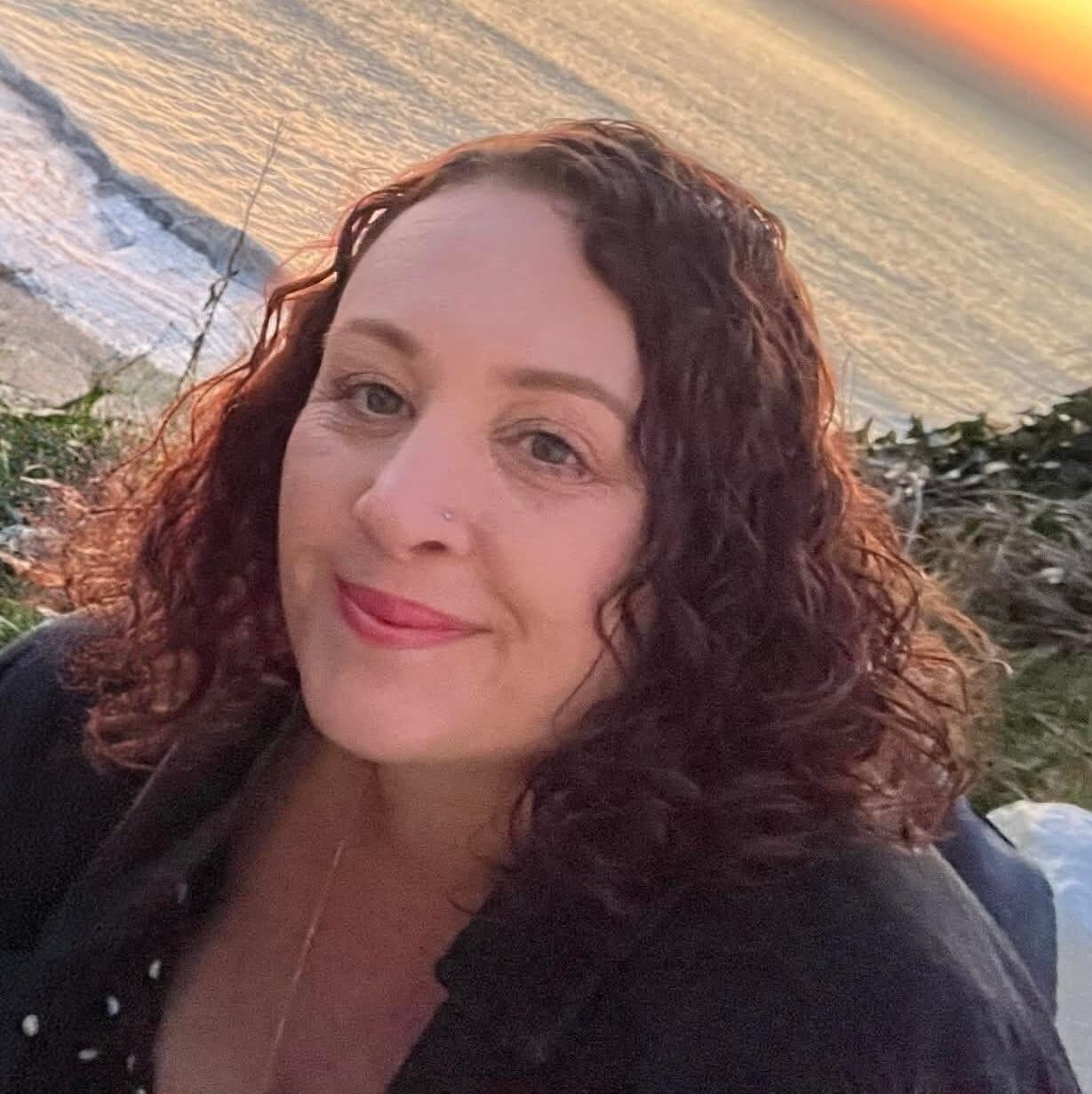 Nikita from Holistic Beatz smiling on a clifftop, with the sea glowing in a golden sunset behind her