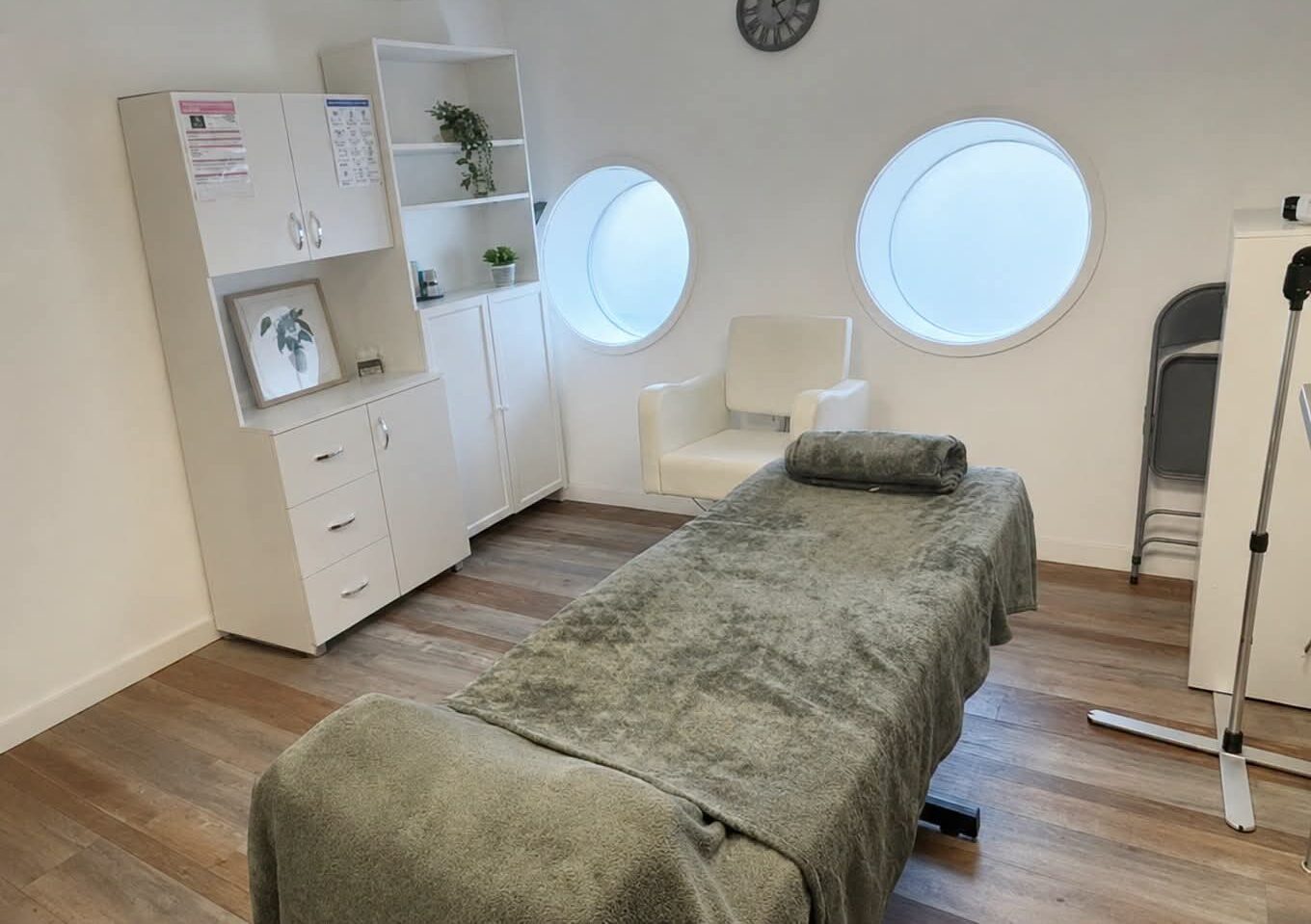 A bright treatment room with a massage couch, round porthole windows and white cabinets