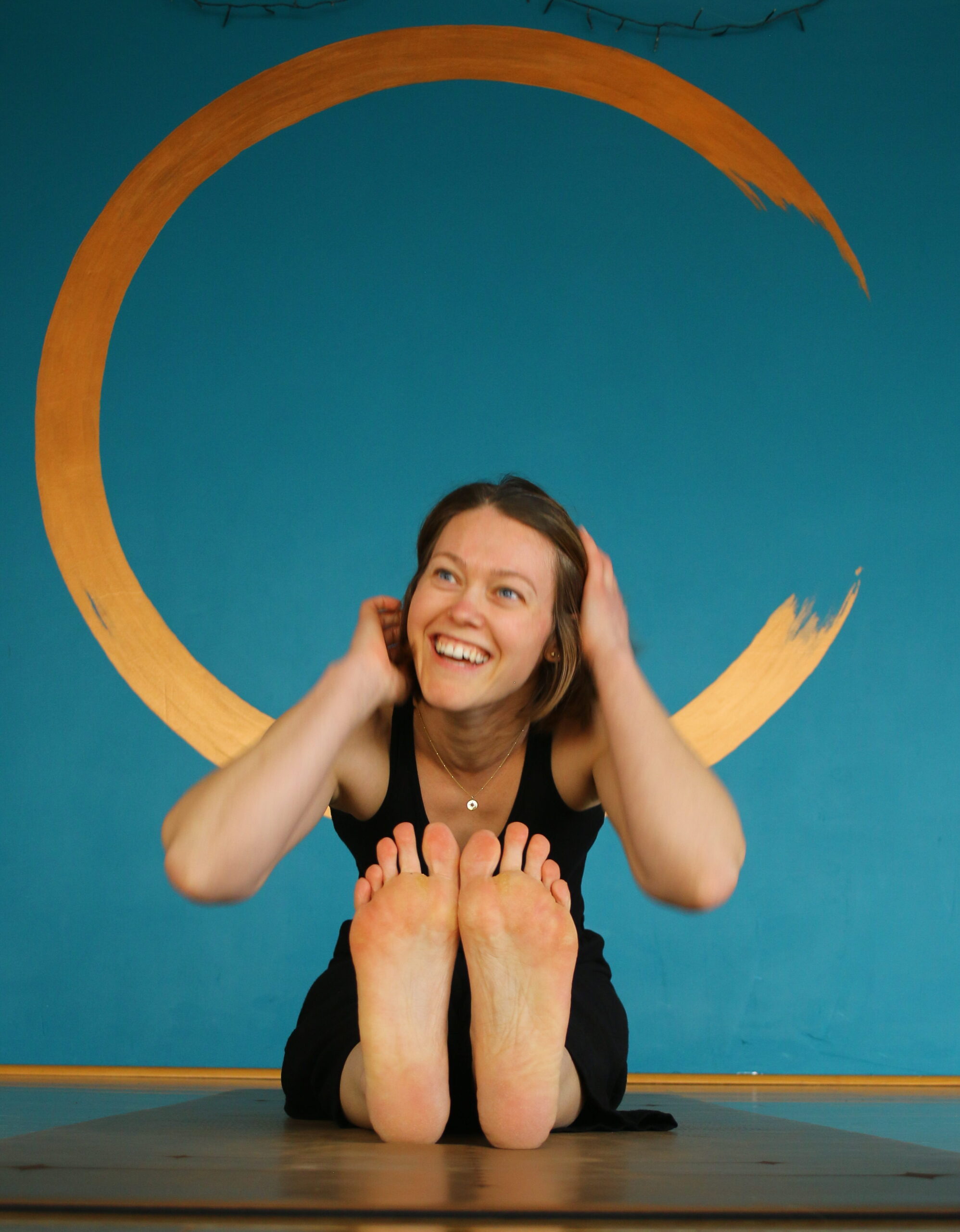 Rachel from Rock and Root Yoga sitting on a wooden studio floor, smiling, with a golden circle painted on the teal wall behind her