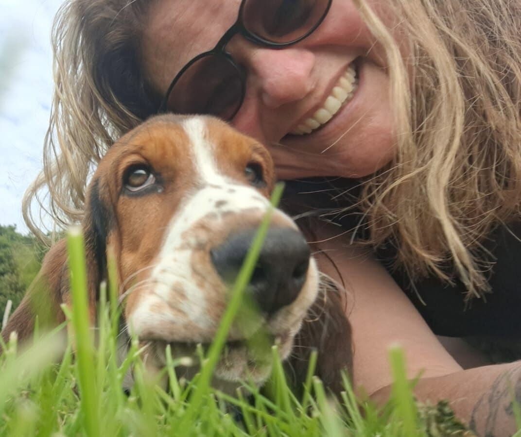 Rachel from Sentu Complementary Therapies smiling in sunglasses, lying in the grass cheek to cheek with her basset hound