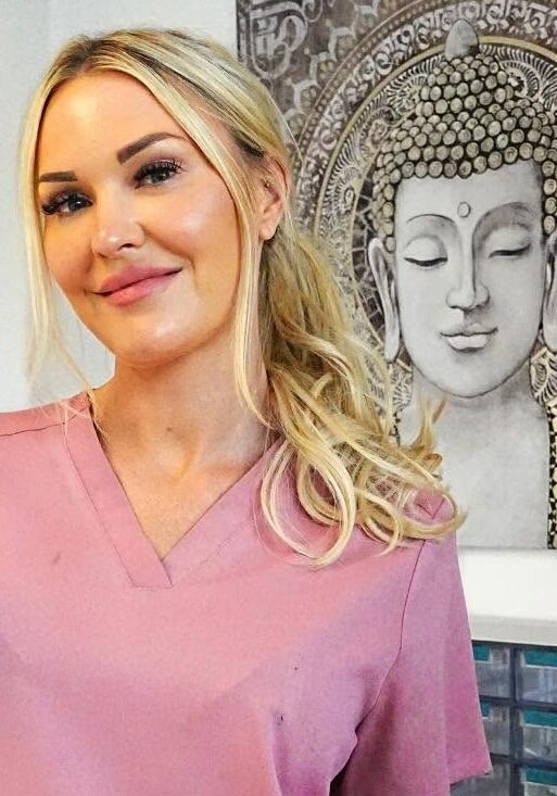 Sophie from Azure Aesthetics Wellbeing smiling in pink scrubs in her treatment room
