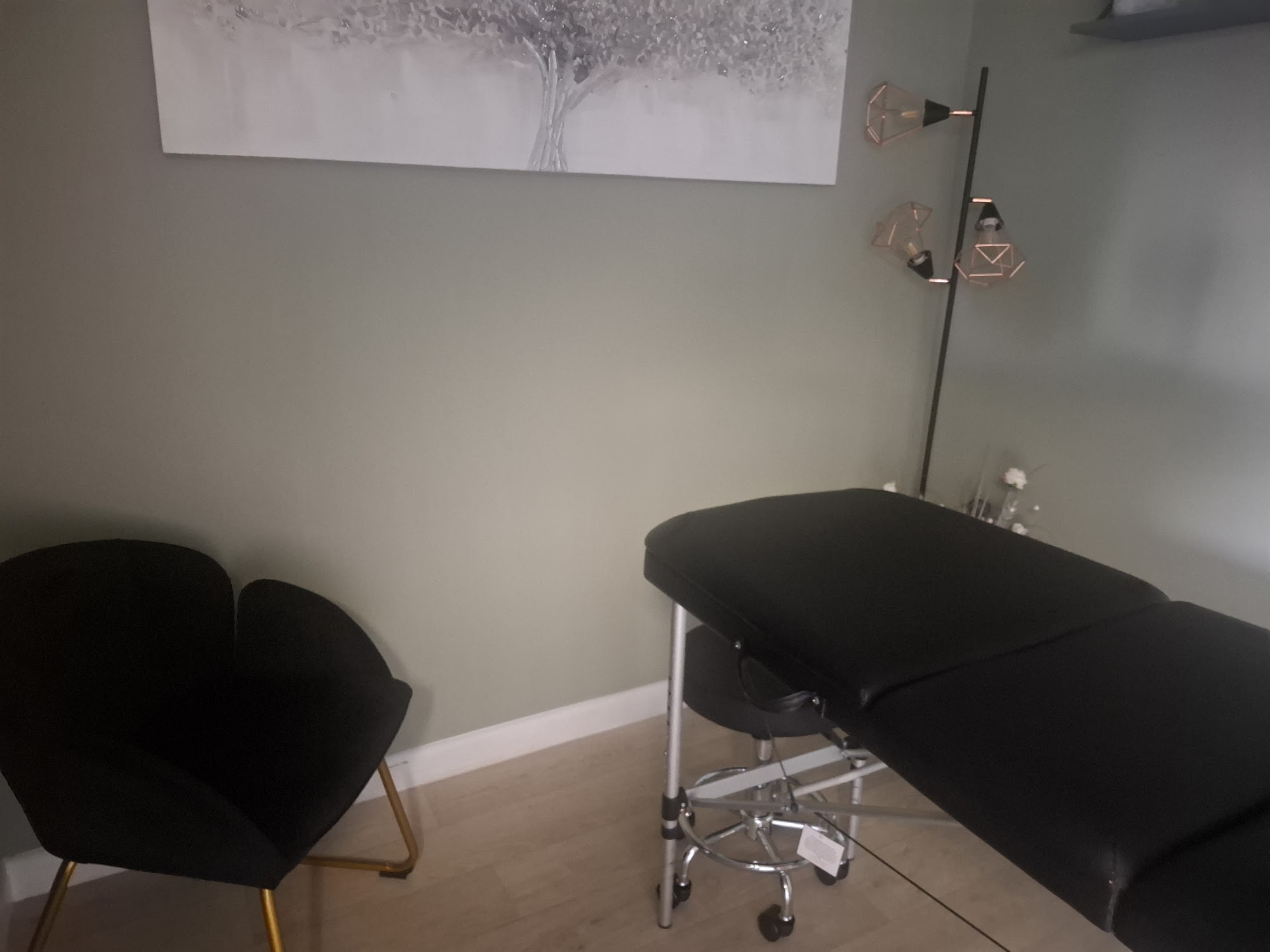 A treatment room with sage green walls, a black massage couch and a black armchair