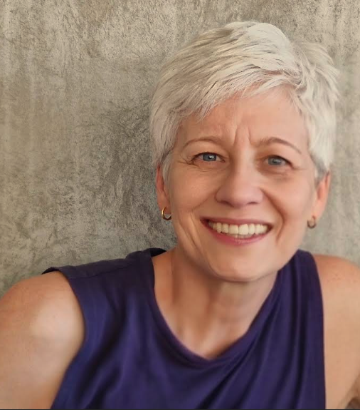 Venita from Yoga with Venita smiling, with short silver hair, wearing a purple sleeveless top against a textured grey wall